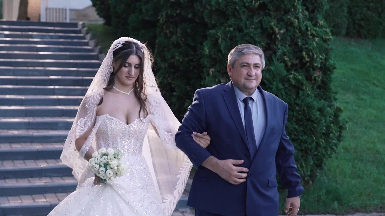 Невеста в длинной фате идёт под руку с седовласым мужчиной в синем костюме; позади лестница и зелень.